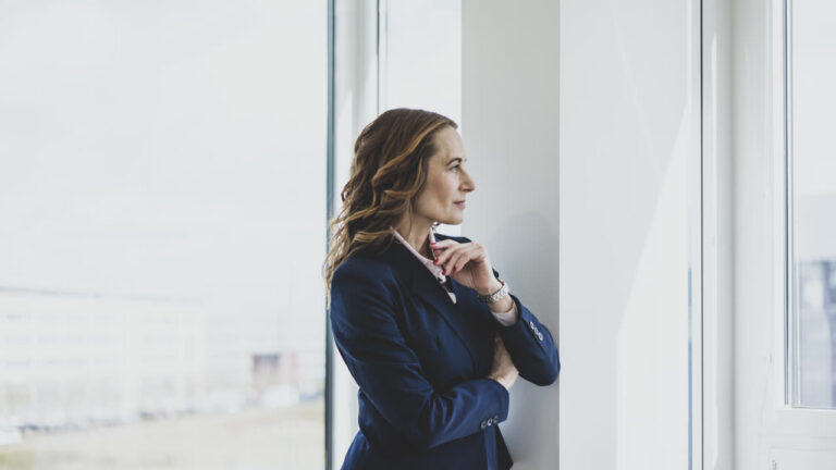 A woman standing by an office window, pausing to look out during the workday
