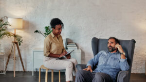 A counselor with a clipboard listening to a man seated in an armchair during a therapy session