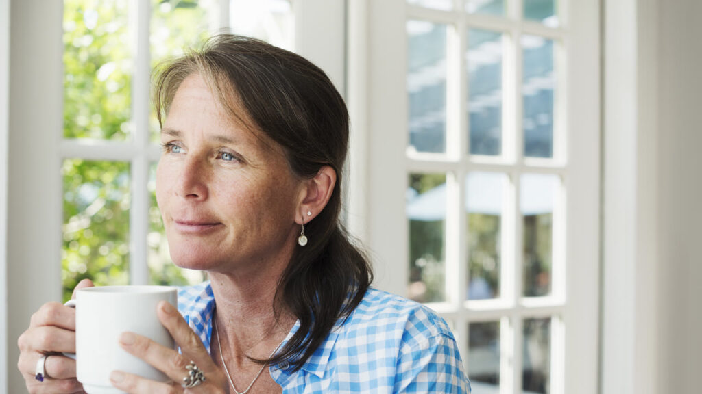 A woman holding a mug of tea and looking out a sunlit window, deep in thought
